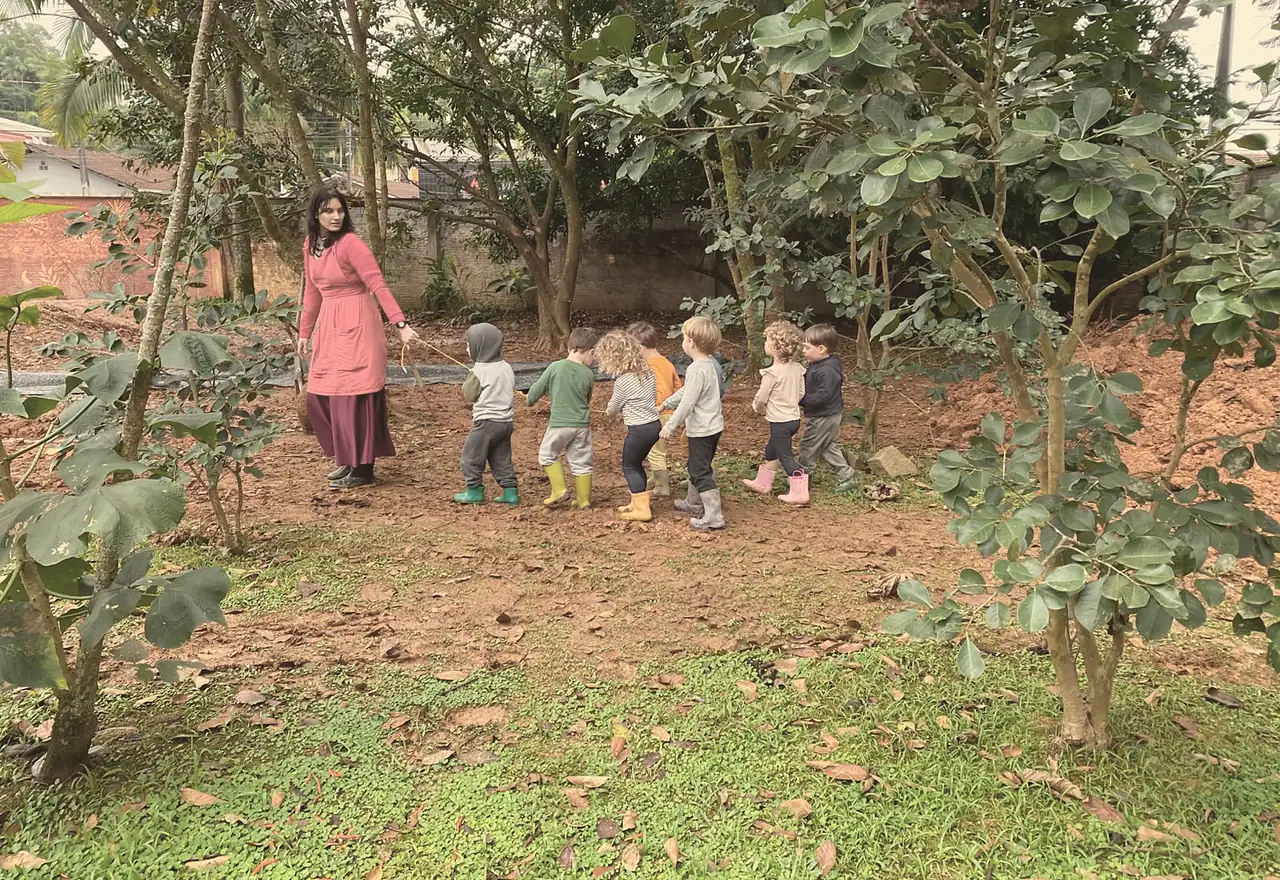 Professora conduz uma fila de crianças, vistas de costas e de botas de chuva, segurando uma corda entre as árvores do jardim