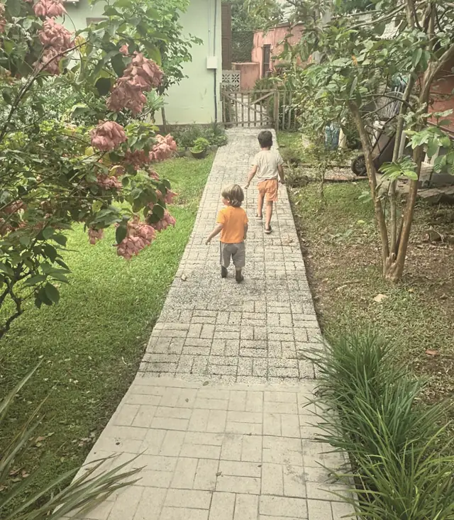 Duas crianças, de costas, caminhando por uma trilha de pedras entre a grama e arbustos floridos