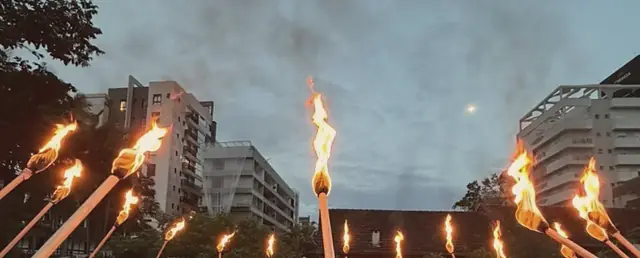 Tochas acesas erguidas em roda ao entardecer, durante uma festa da escola, com céu nublado e a lua ao fundo