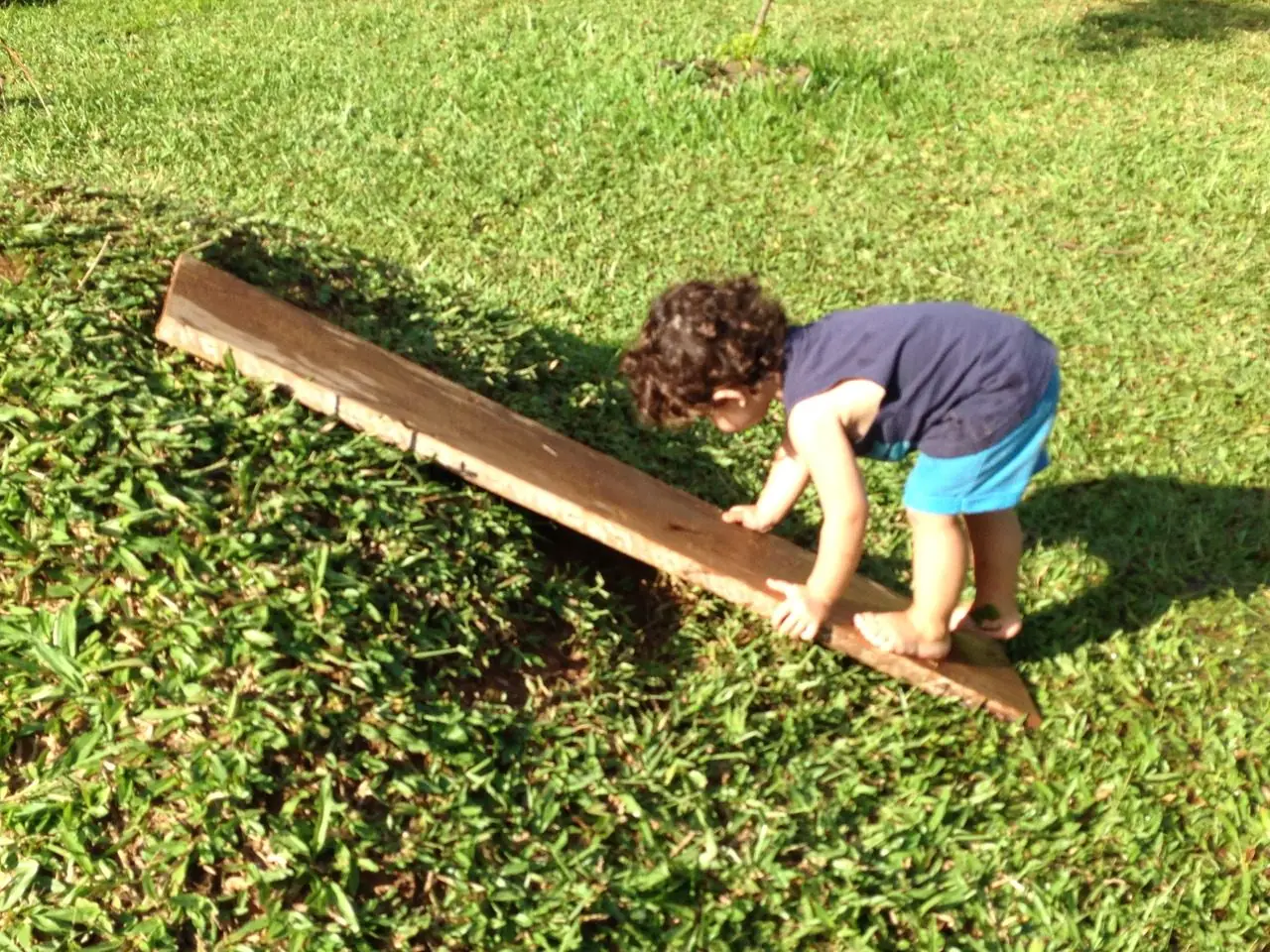 Criança descalça, com o rosto voltado para baixo, apoiando as mãos numa tábua de madeira inclinada sobre a grama ensolarada