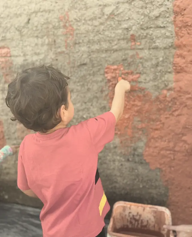 Criança de costas pintando um muro com tinta de terra avermelhada, ao lado de um balde de tinta